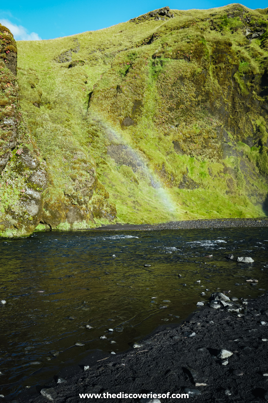 A rainbow arching through the spray over the dark Skógá river below Skógafoss, with green slopes behind