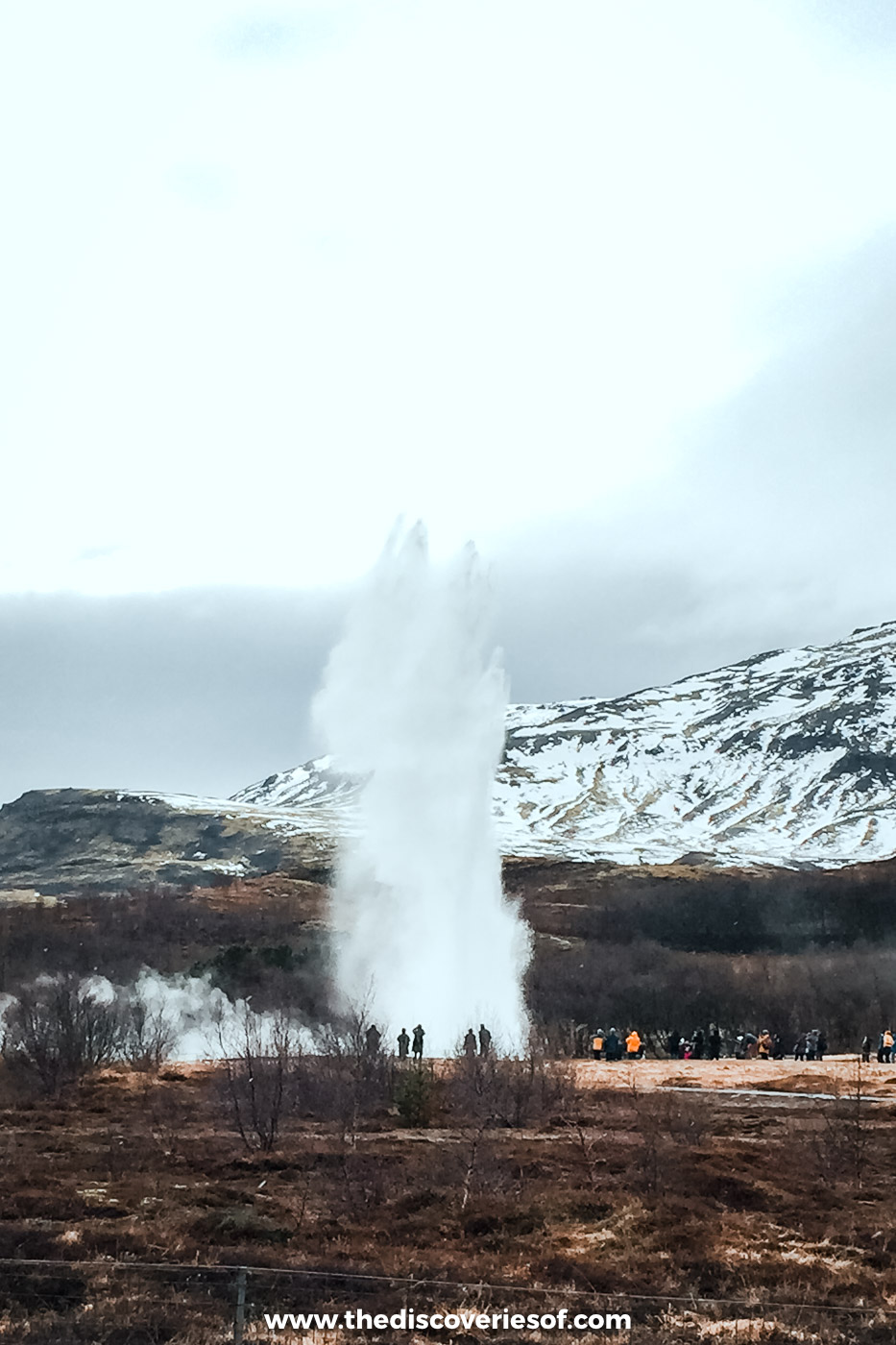 Strokkur geyser erupting in a tall white column below snow-streaked hills, with visitors along the rope, Haukadalur, Iceland