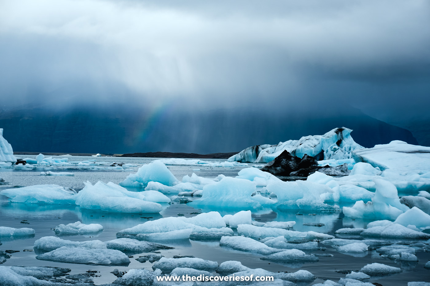 A dark rain squall sweeping over blue and white icebergs at Jökulsárlón, with a faint rainbow beneath the cloud