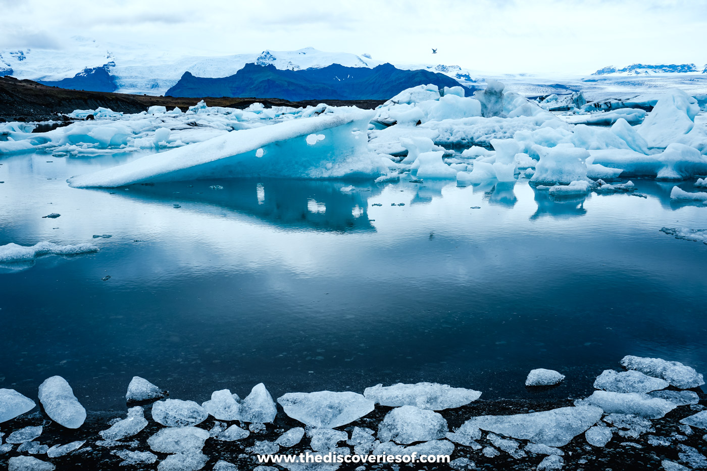 A pale blue iceberg reflected in the still water of Jökulsárlón with mountains behind