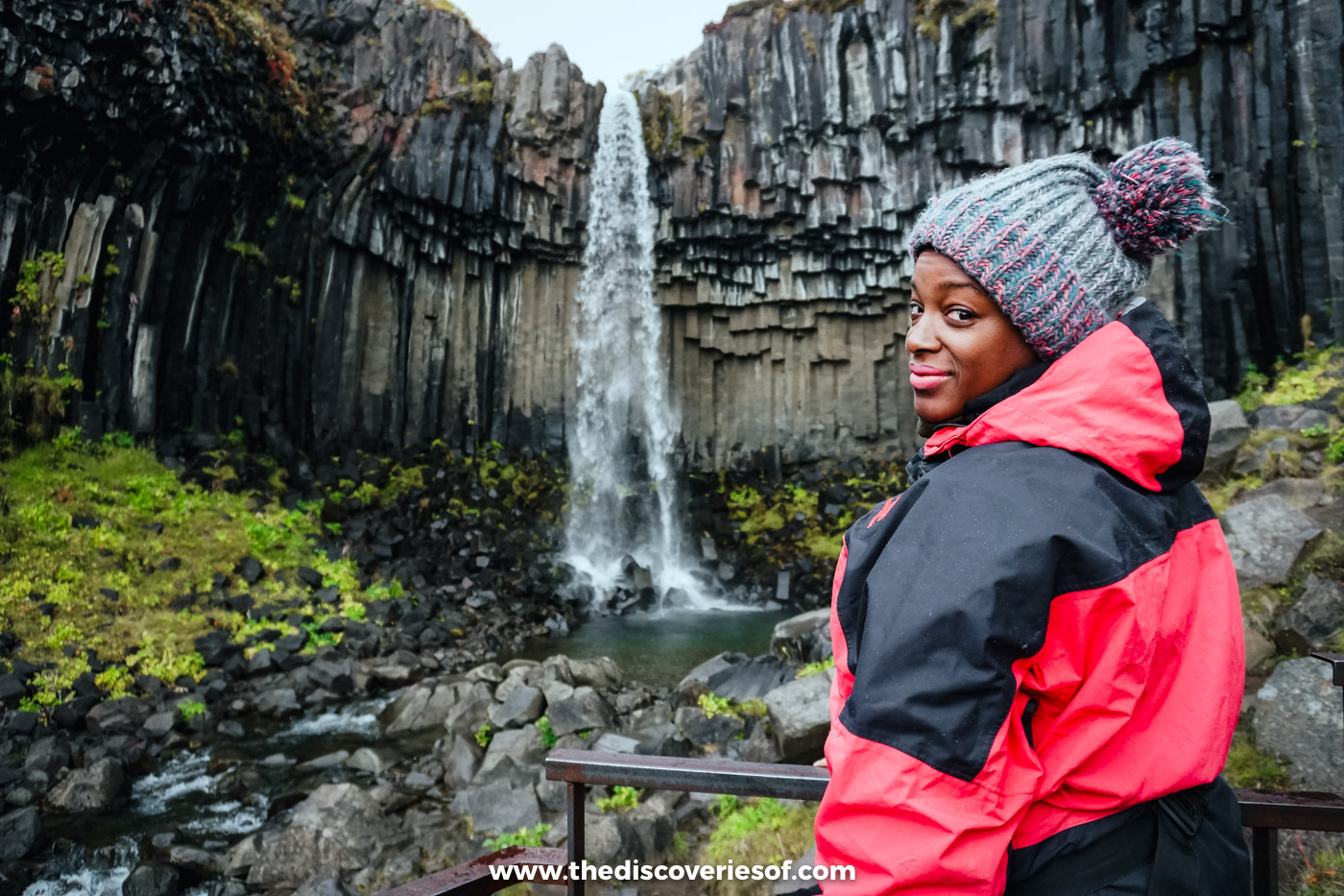 Julianna in a red and black jacket and grey bobble hat smiling in front of Svartifoss and its basalt columns