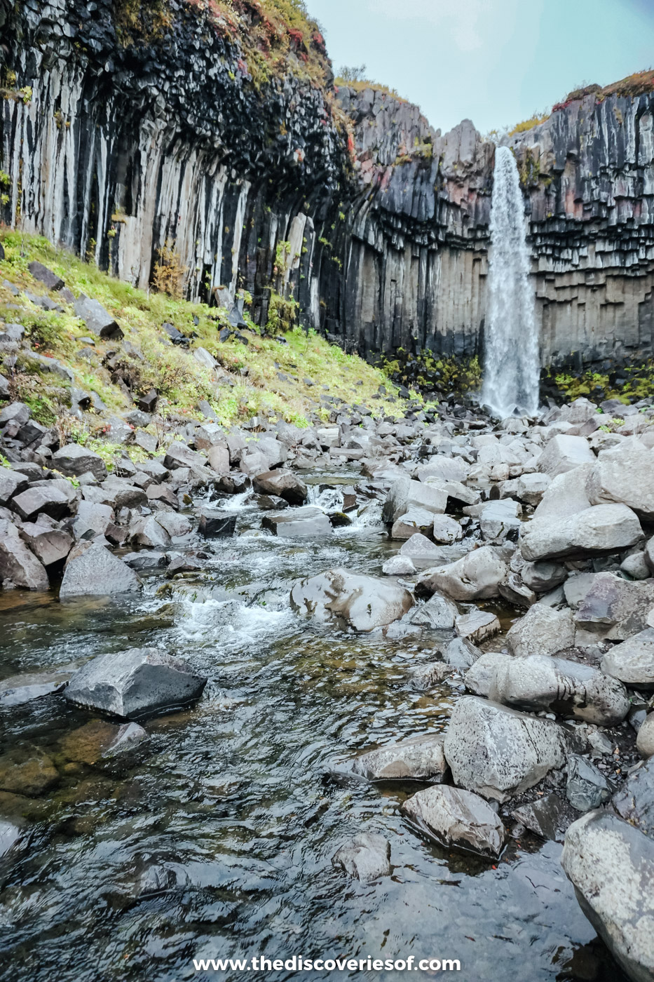 Stream running over grey boulders and fallen basalt below Svartifoss and its dark column cliff in Skaftafell