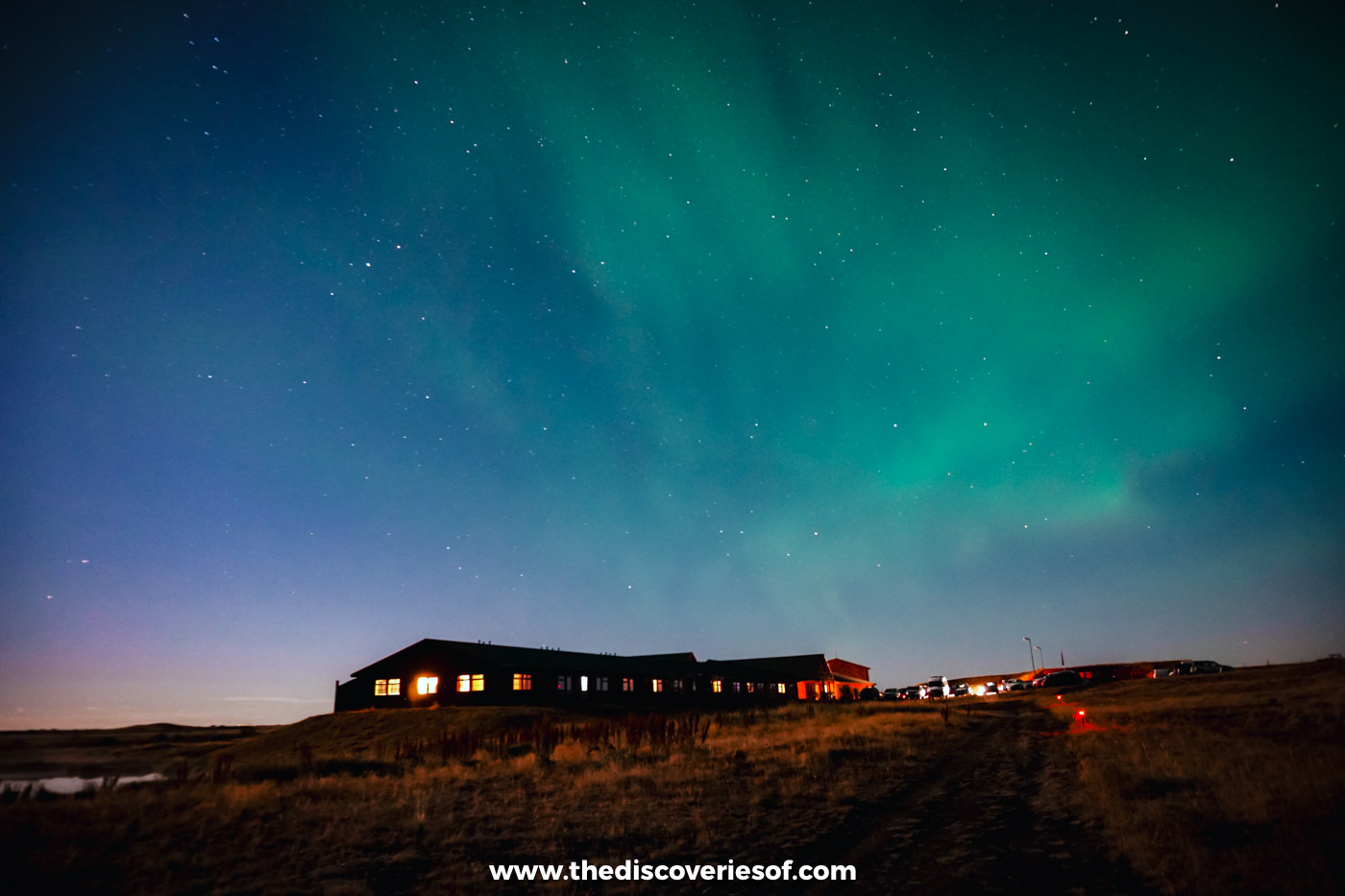Green Northern Lights spreading across a starry sky above a long low lit building in the south Iceland countryside