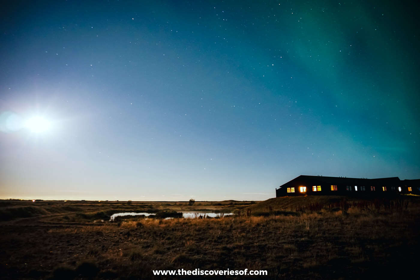 Green aurora spreading across the night sky beside a bright, almost full moon over the low buildings of Hotel Rangá