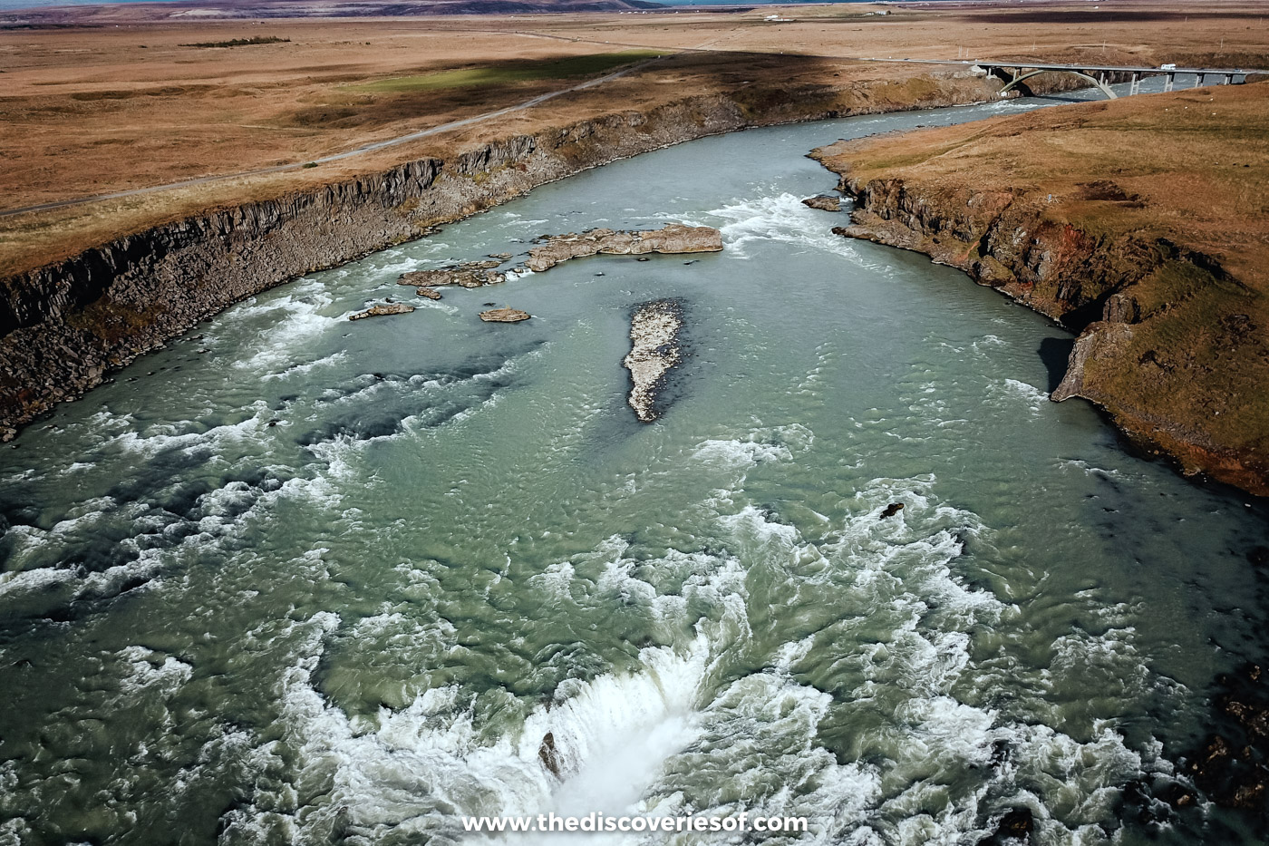 Drone shot over the lip of Urriðafoss looking up the wide grey-green Þjórsá towards distant hills