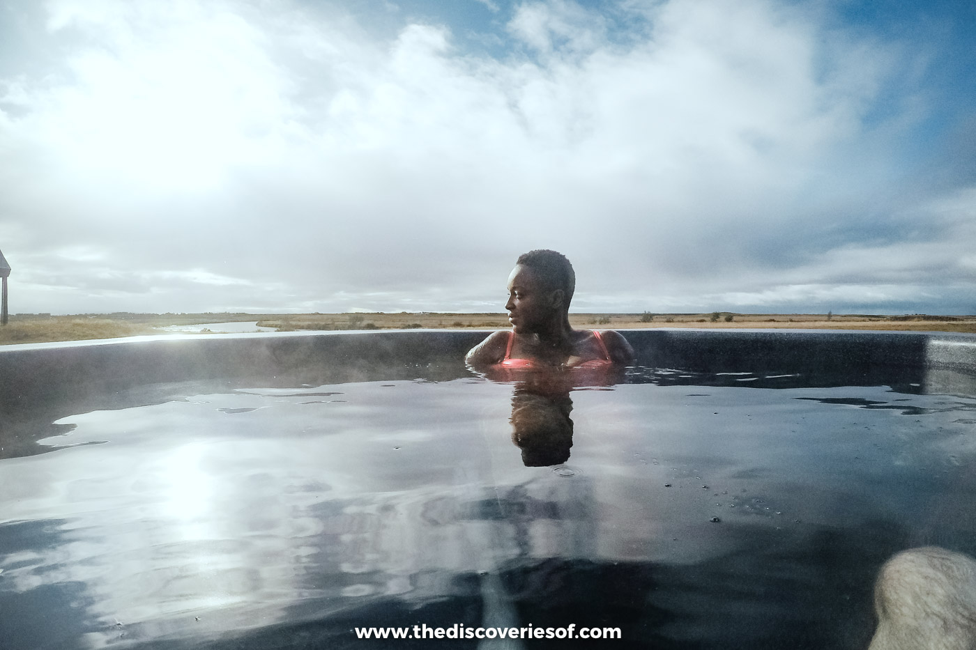 Julianna relaxing in an outdoor hot tub with a flat landscape and cloudy sky behind in South Iceland