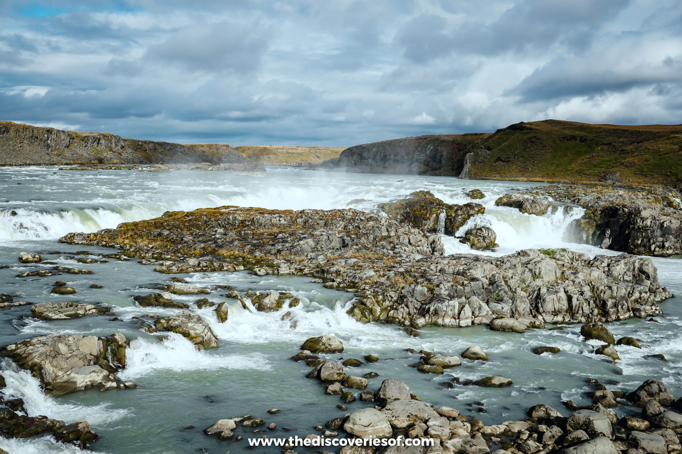 Urriðafoss seen from the west bank, water pouring around a broad rocky ledge with mist and low cliffs behind