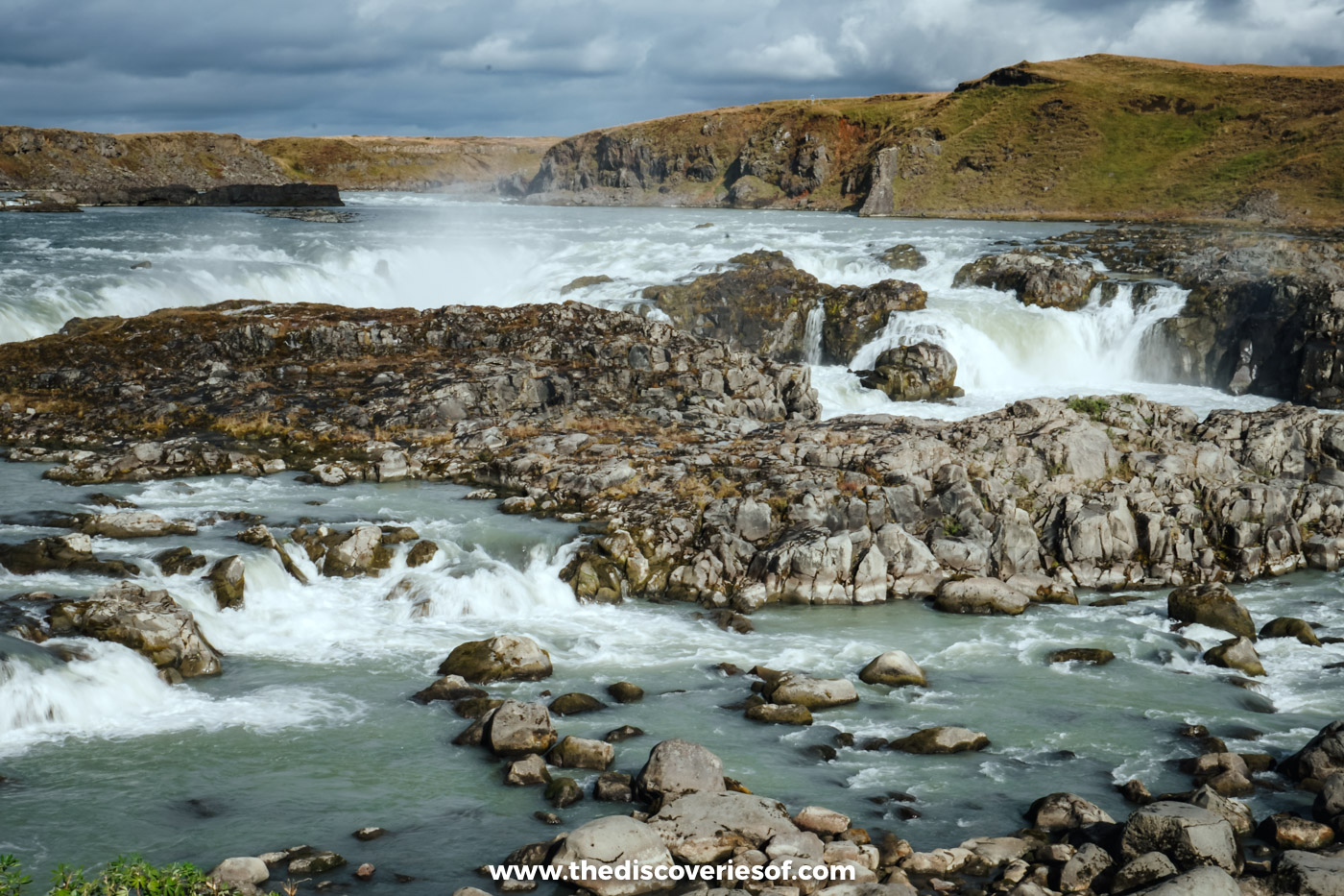 Rapids running between dark rocks and small cascades at Urriðafoss, with the far bank under a cloudy sky