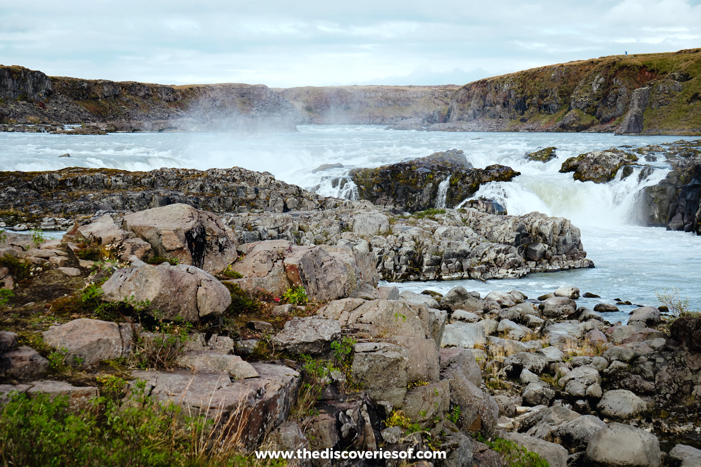 Pink-grey boulders and grass on the bank of the Þjórsá with Urriðafoss and its spray behind