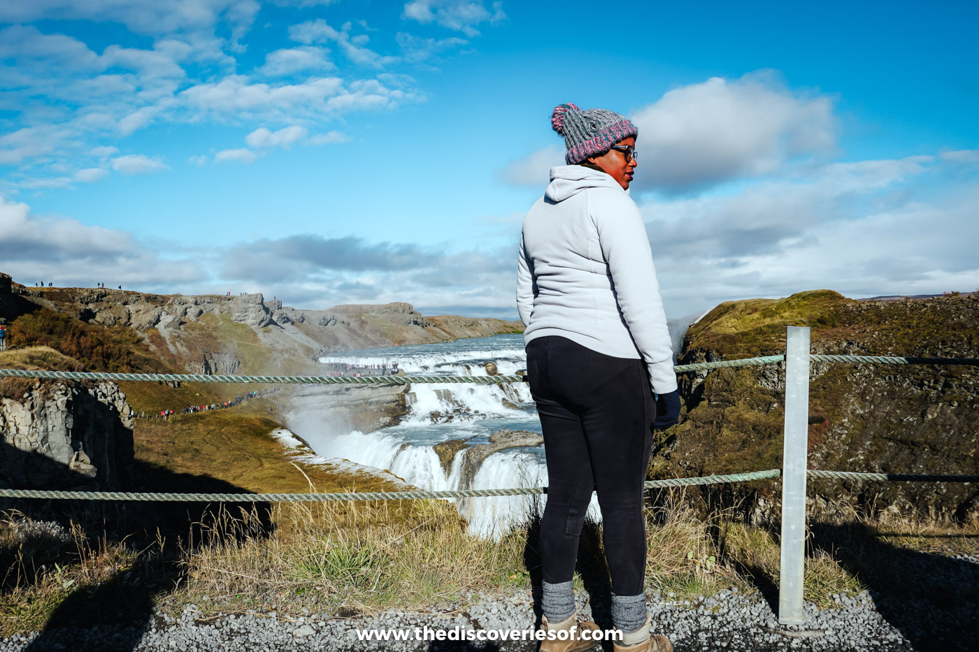 Julianna in a bobble hat and pale hoodie standing at the rope railing above Gullfoss in September