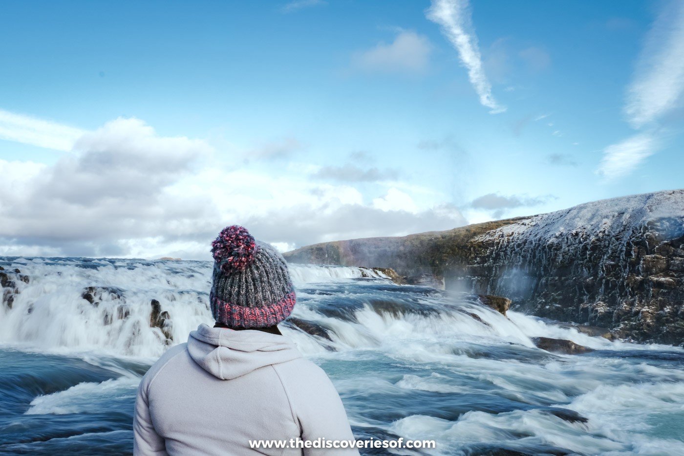 Julianna seen from behind in a bobble hat, standing right beside the rushing first step of Gullfoss