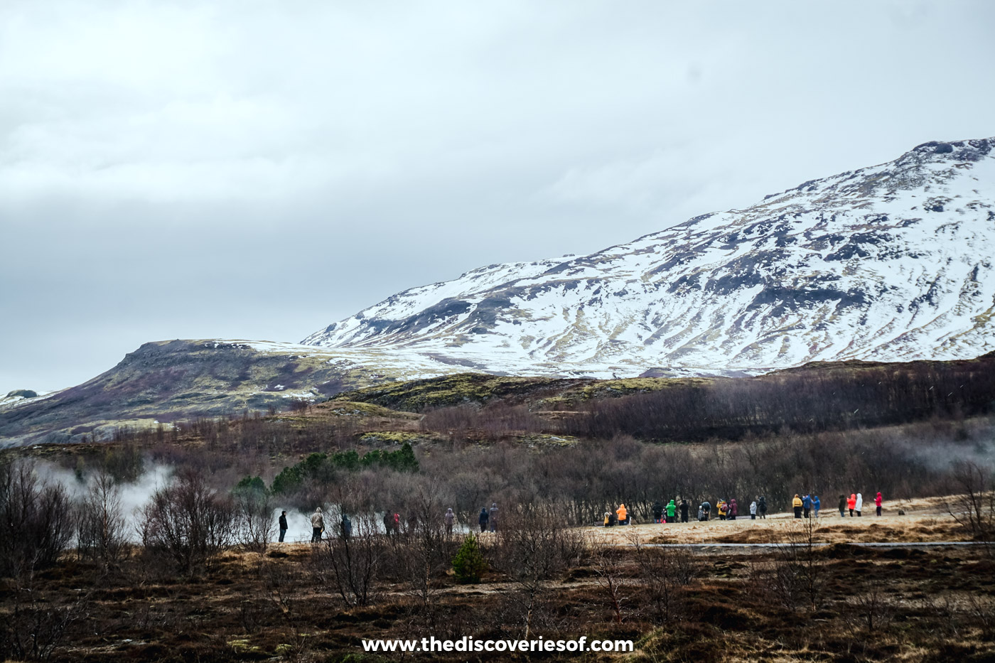 Steam rising from the Geysir geothermal field below a snow-covered mountain, with visitors on the path in early May