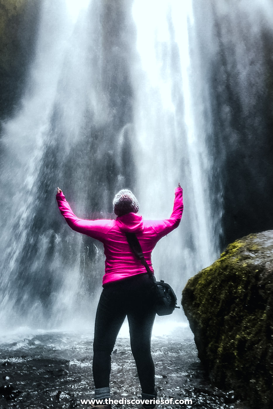 Woman in a pink jacket raising her arms in the spray at the foot of Gljúfrabúi waterfall in its mossy gorge