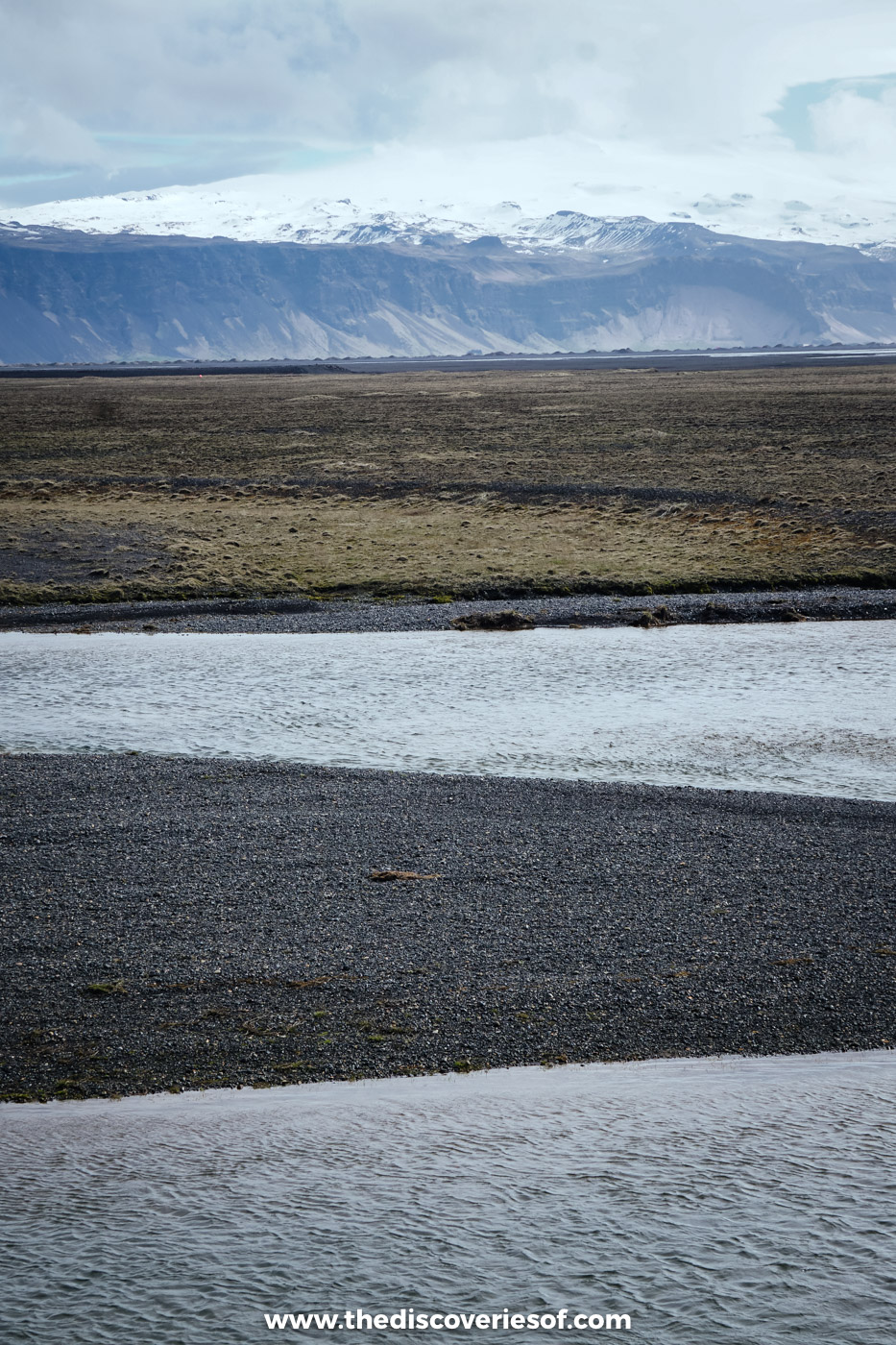 Snow-streaked ridge rising behind a wide plain of black sand and shallow meltwater in south Iceland in early May