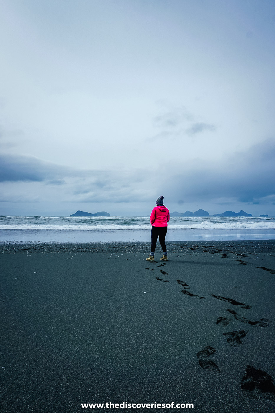 Woman in a pink jacket and bobble hat walking across black sand towards the sea at Landeyjafjara in south Iceland