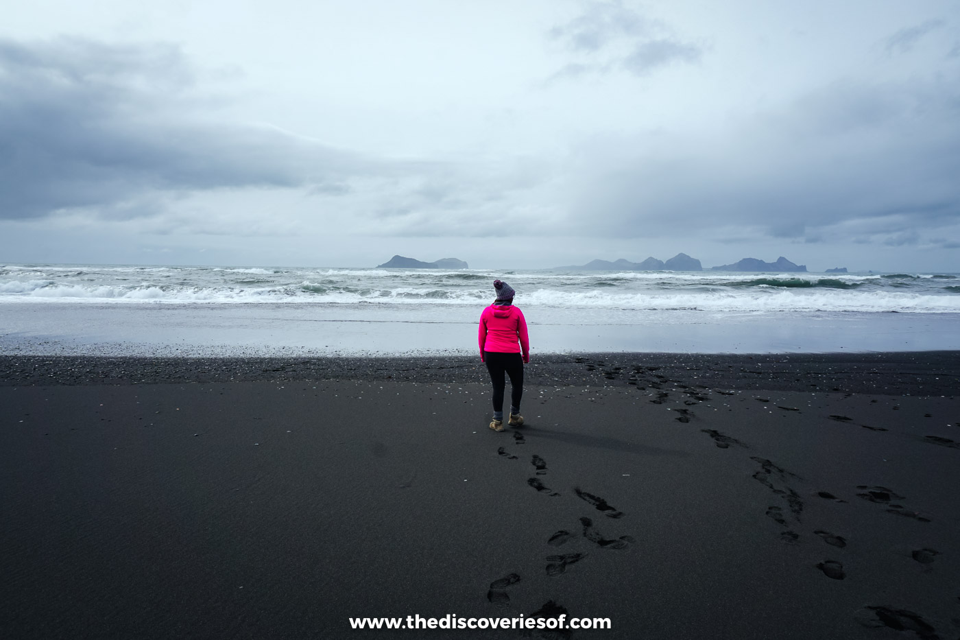 Julianna in a pink jacket walking across black sand towards the surf at Landeyjafjara with the Westman Islands beyond