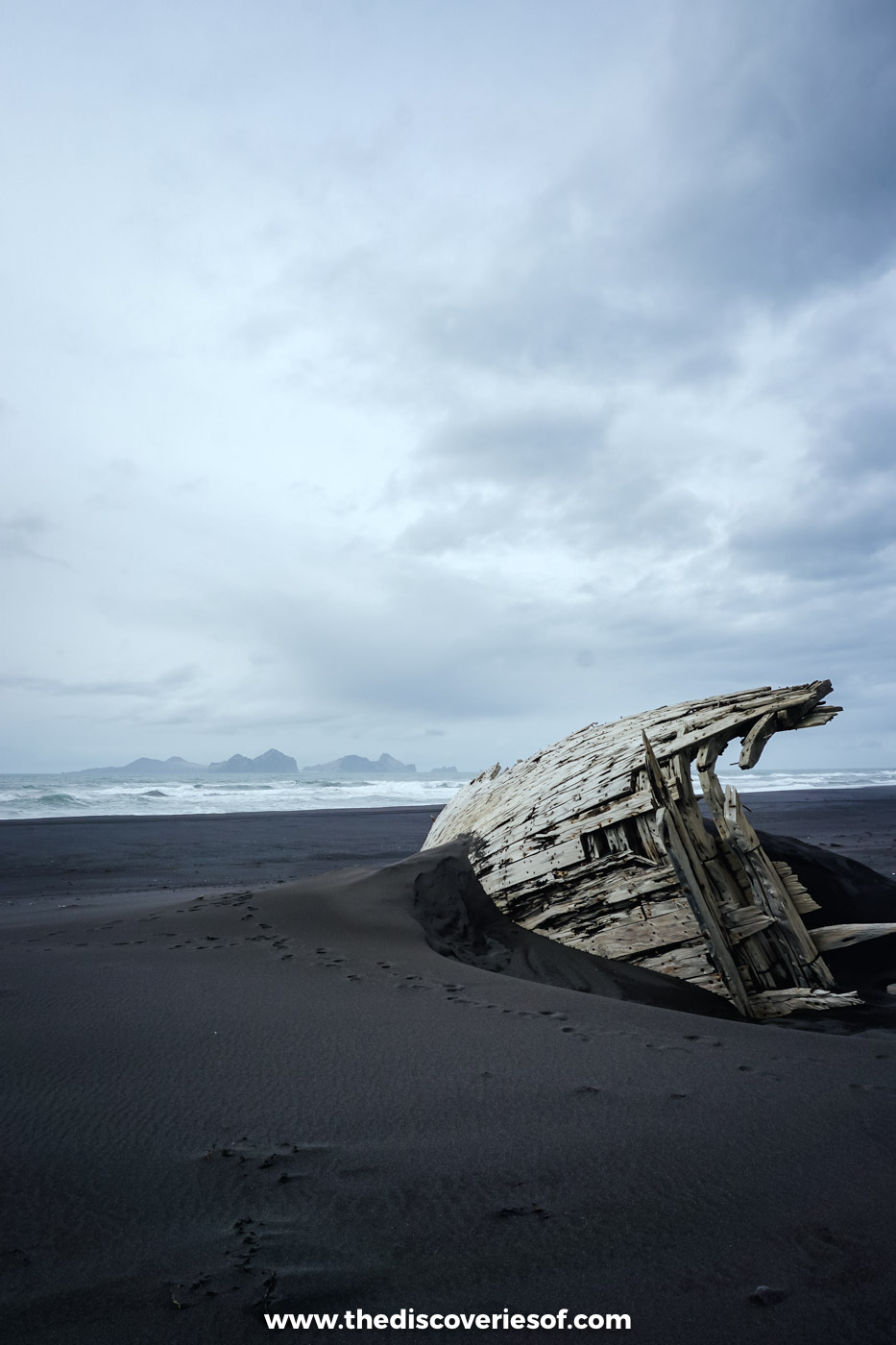 The weathered timbers of an old wooden boat half buried in black sand at Landeyjafjara under a grey sky