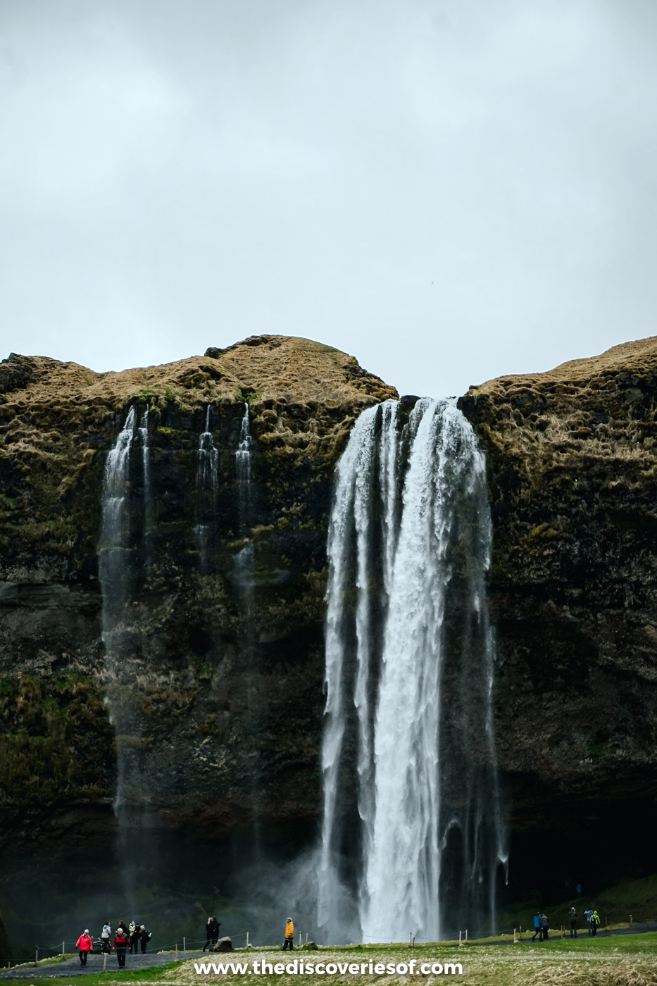 Seljalandsfoss dropping from a dark mossy cliff into its pool, with visitors on the path at the base