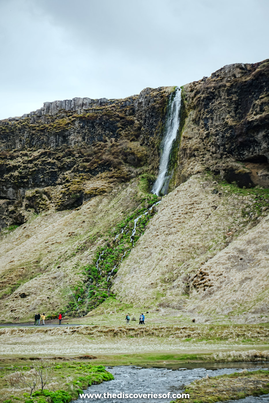 A thin waterfall spilling down a mossy cliff beside Seljalandsfoss, with walkers on the path below
