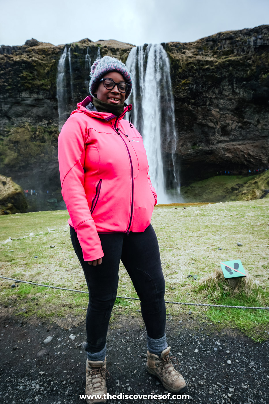 Julianna in a pink jacket and grey bobble hat standing on the grass in front of Seljalandsfoss waterfall