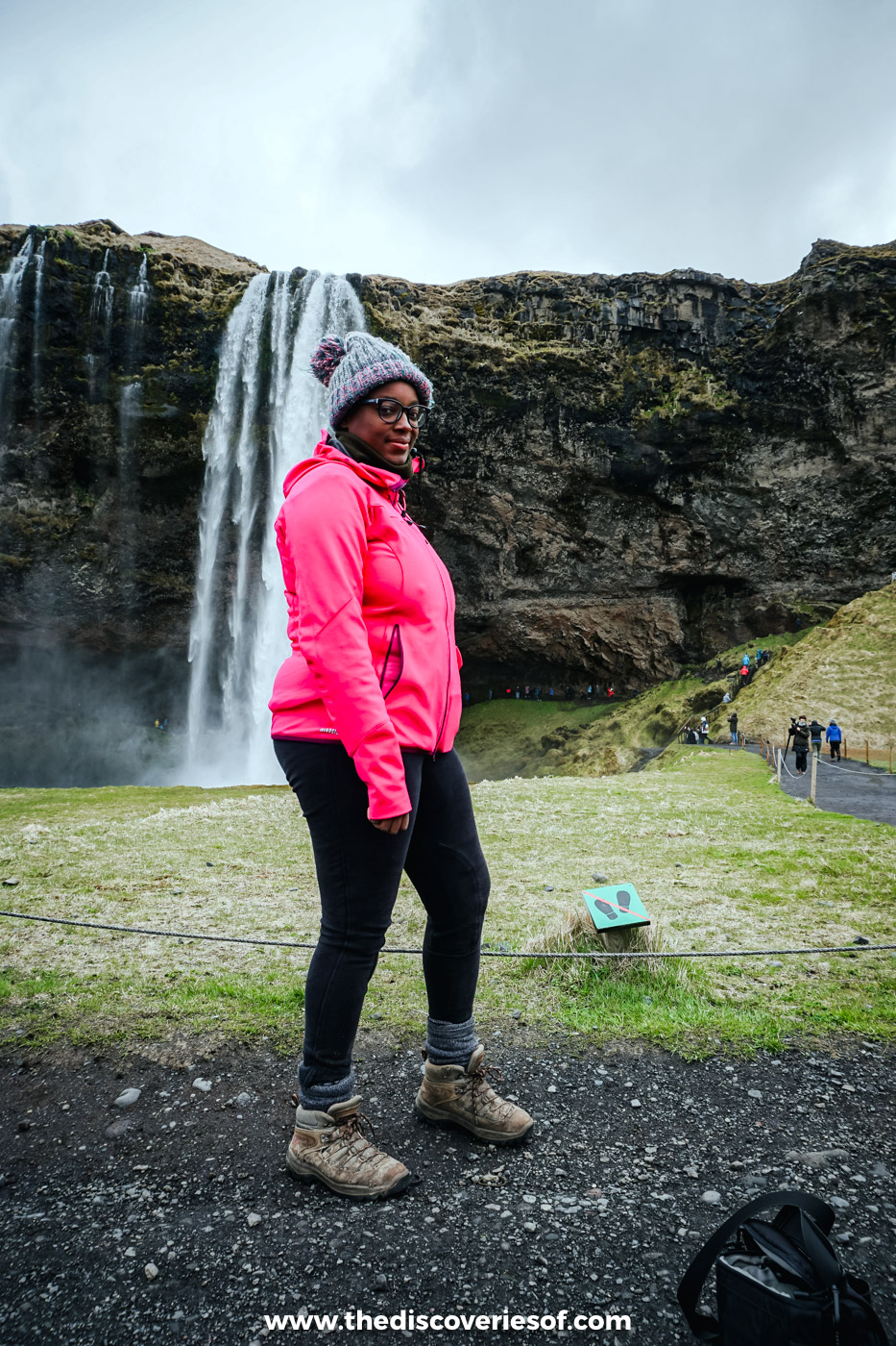 Julianna in a pink jacket and grey bobble hat standing in front of Seljalandsfoss on a grey day in May