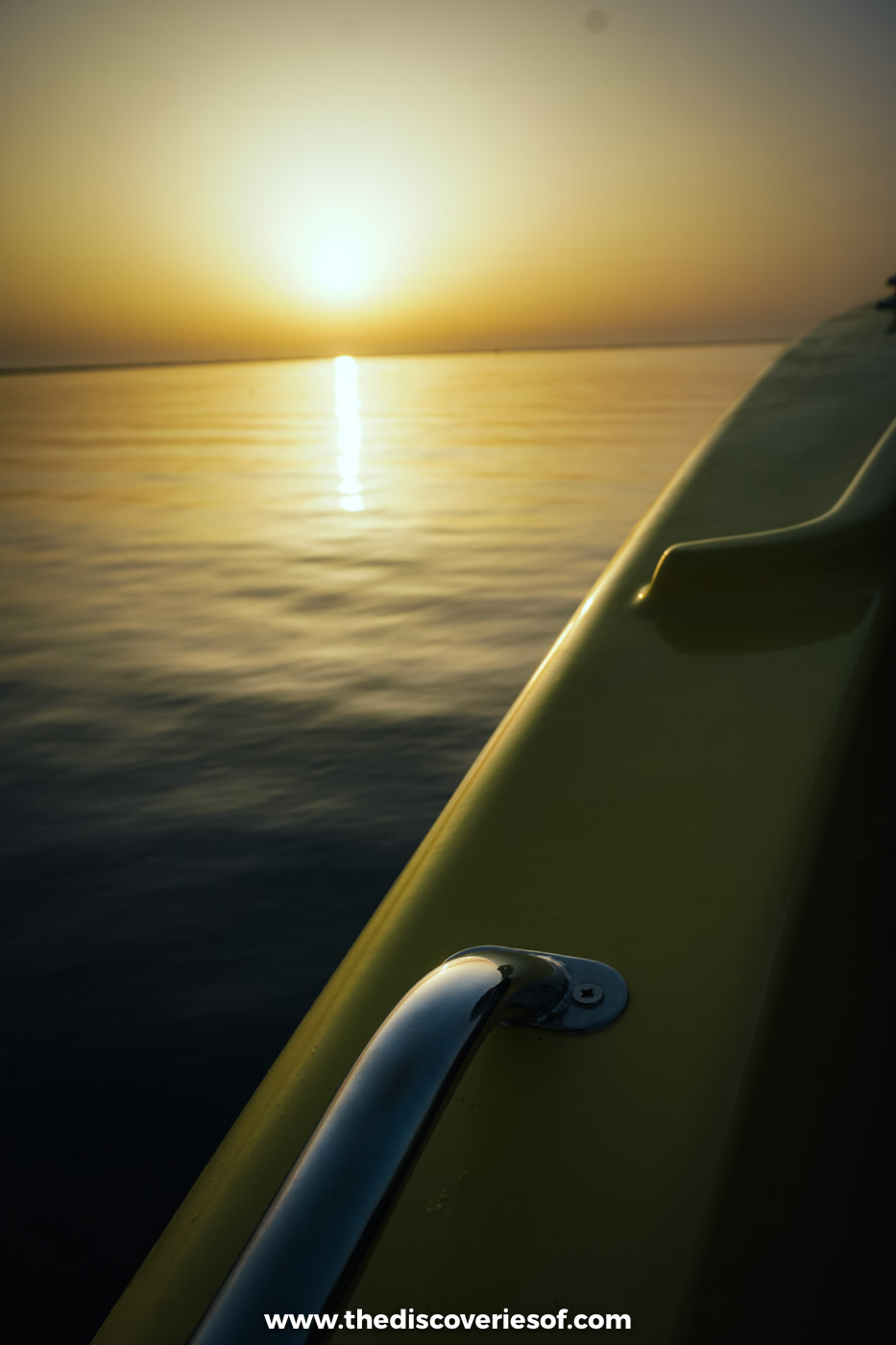 The sun setting over calm golden water, seen past the rail and hull of a boat on the trip to Ilha Deserta