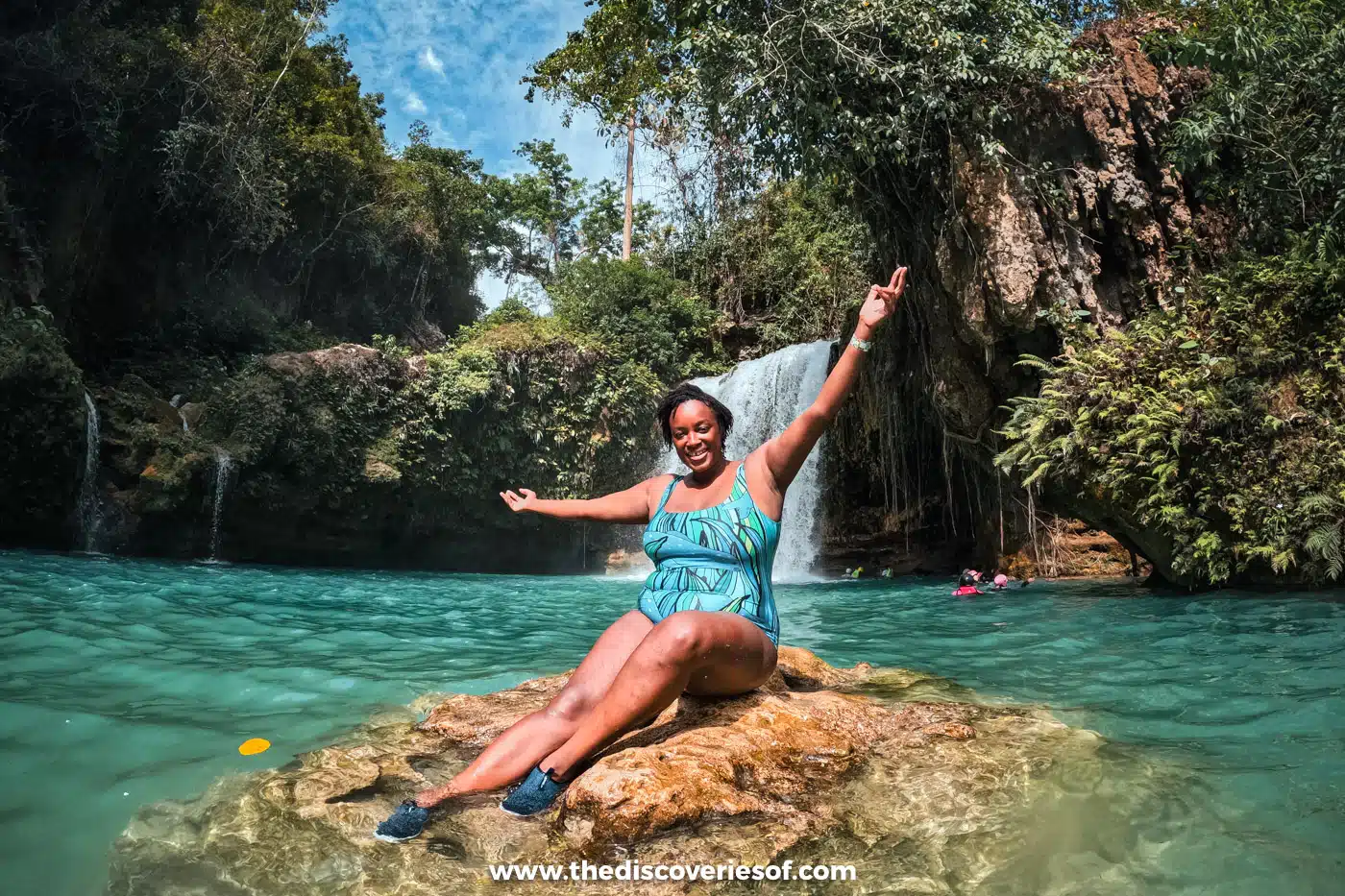 Julianna in a helmet and life jacket sitting on a rock in the turquoise pool at Kawasan Falls, Cebu
