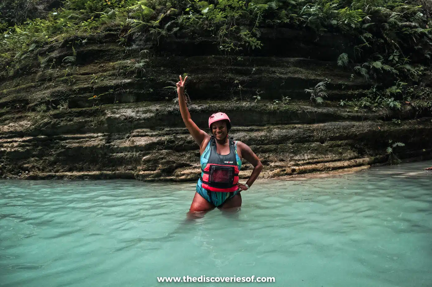 Julianna in a pink helmet wading through turquoise water below layered rock walls in the canyon near Kawasan Falls, Cebu