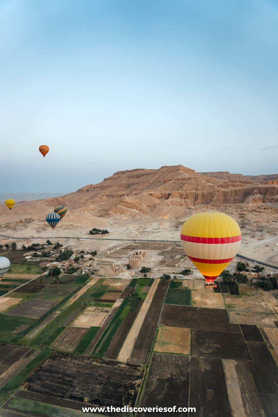 A colourful hot air balloon floating above green farmland on the West Bank of Luxor towards the hills