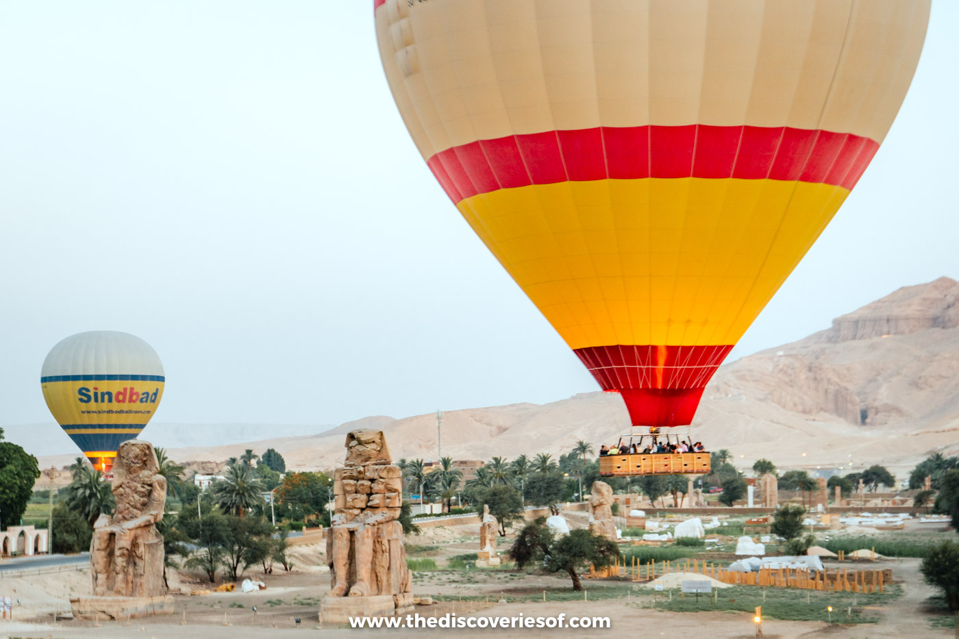 A red and yellow hot air balloon drifting low over the two Colossi of Memnon on Luxor's West Bank