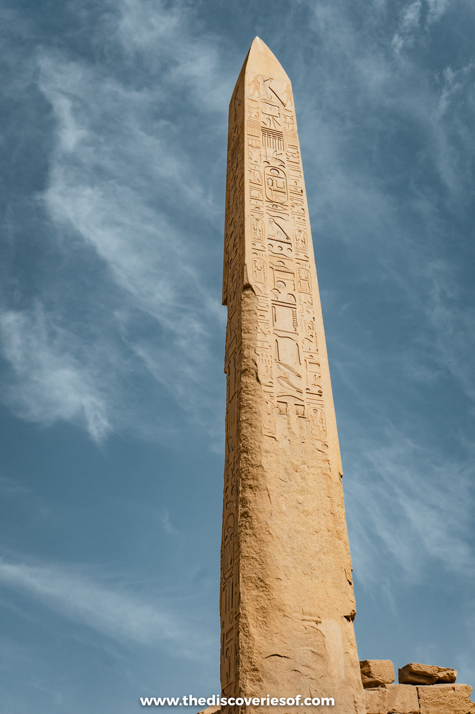 A tall granite obelisk carved with hieroglyphs rising against a blue sky streaked with cloud at Karnak