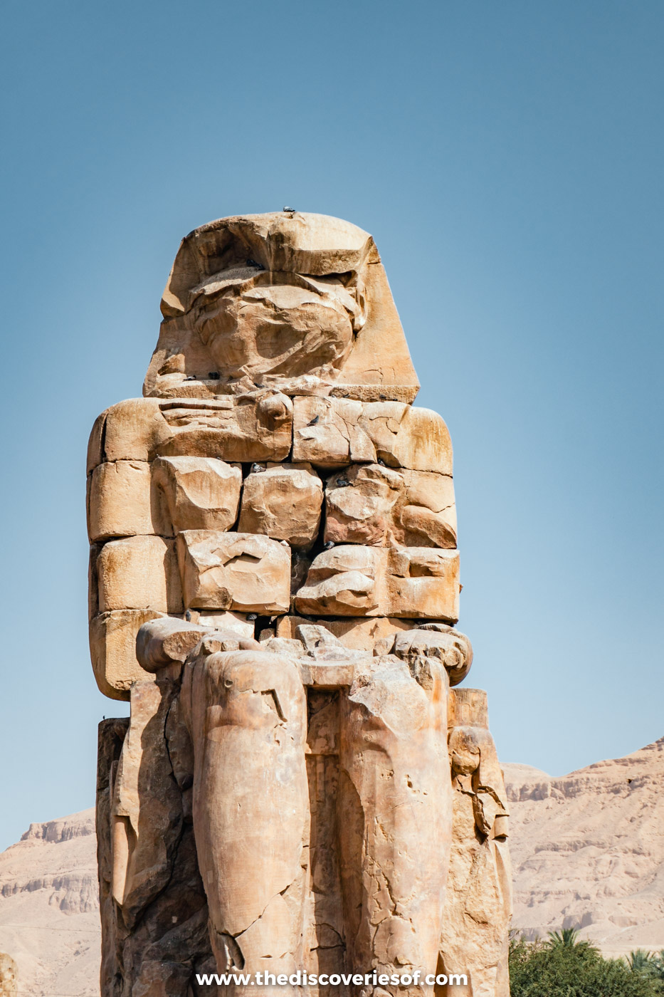Weathered seated colossus of Amenhotep III, rebuilt in stone blocks, against a blue sky at the Colossi of Memnon