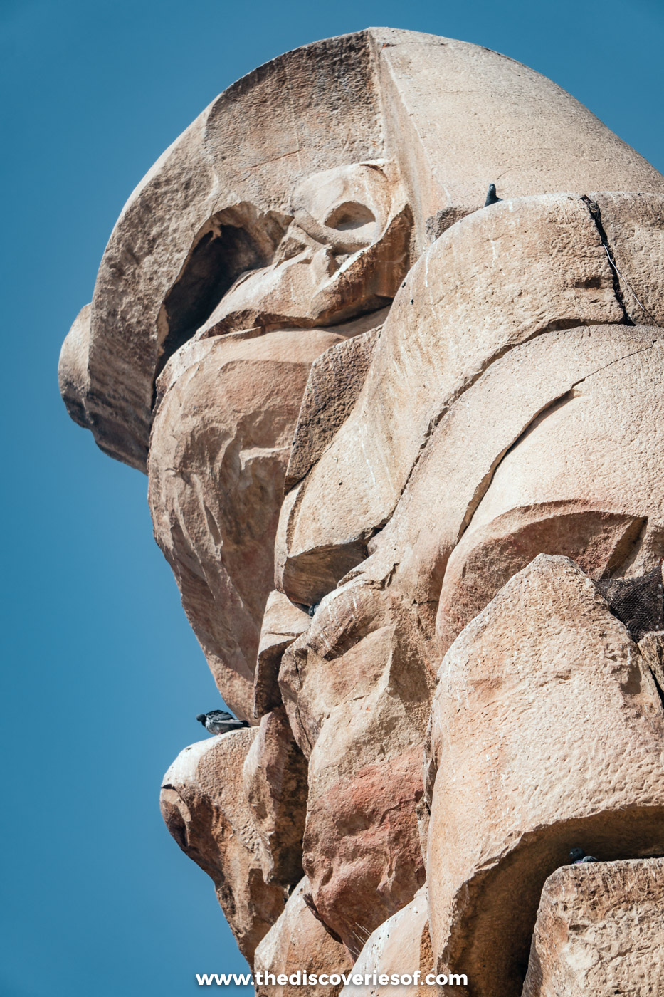 The weathered stone head and shoulders of one of the Colossi of Memnon against a deep blue sky, Luxor