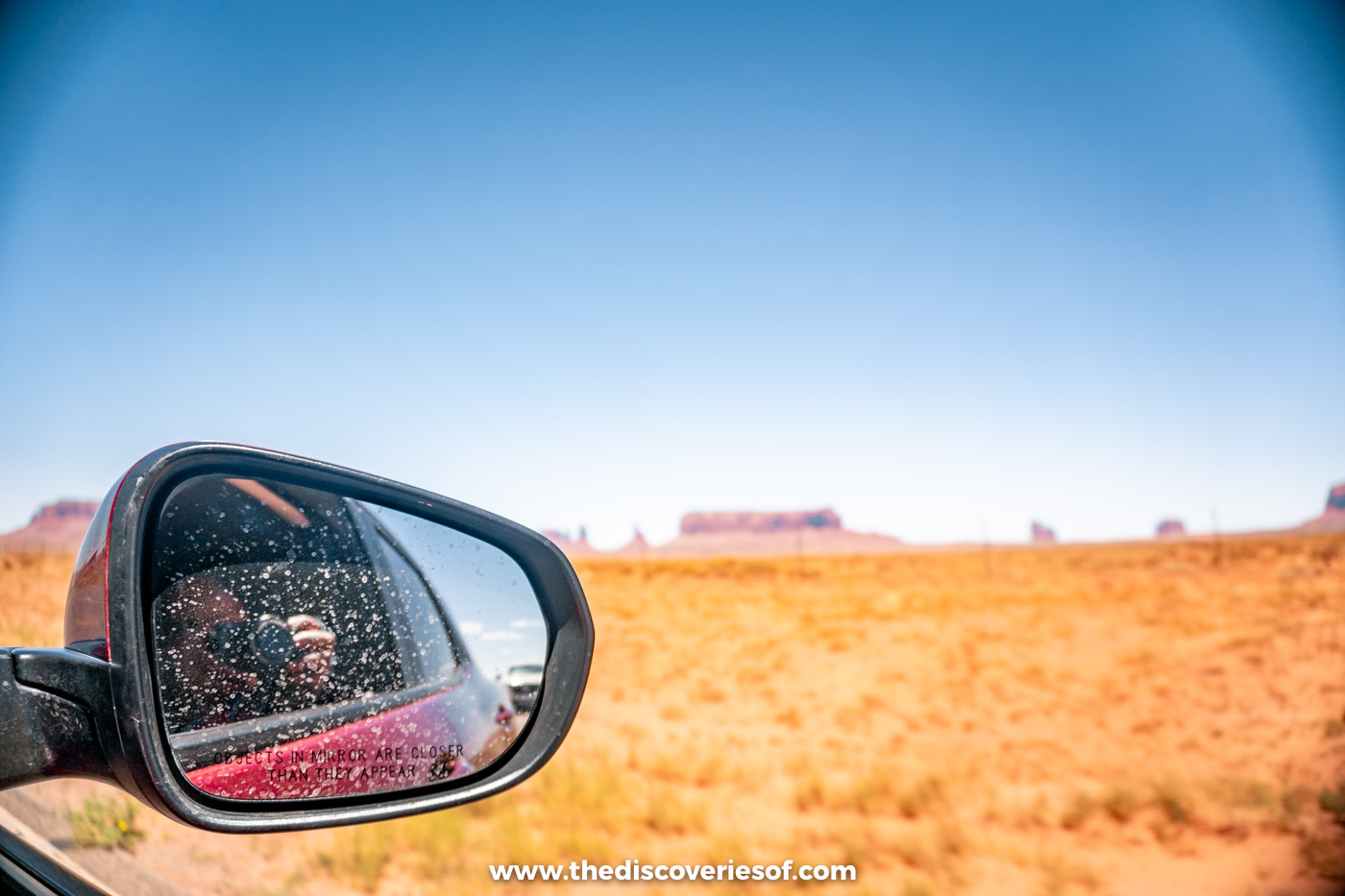 Car wing mirror and grassy desert with distant buttes on the drive into Monument Valley
