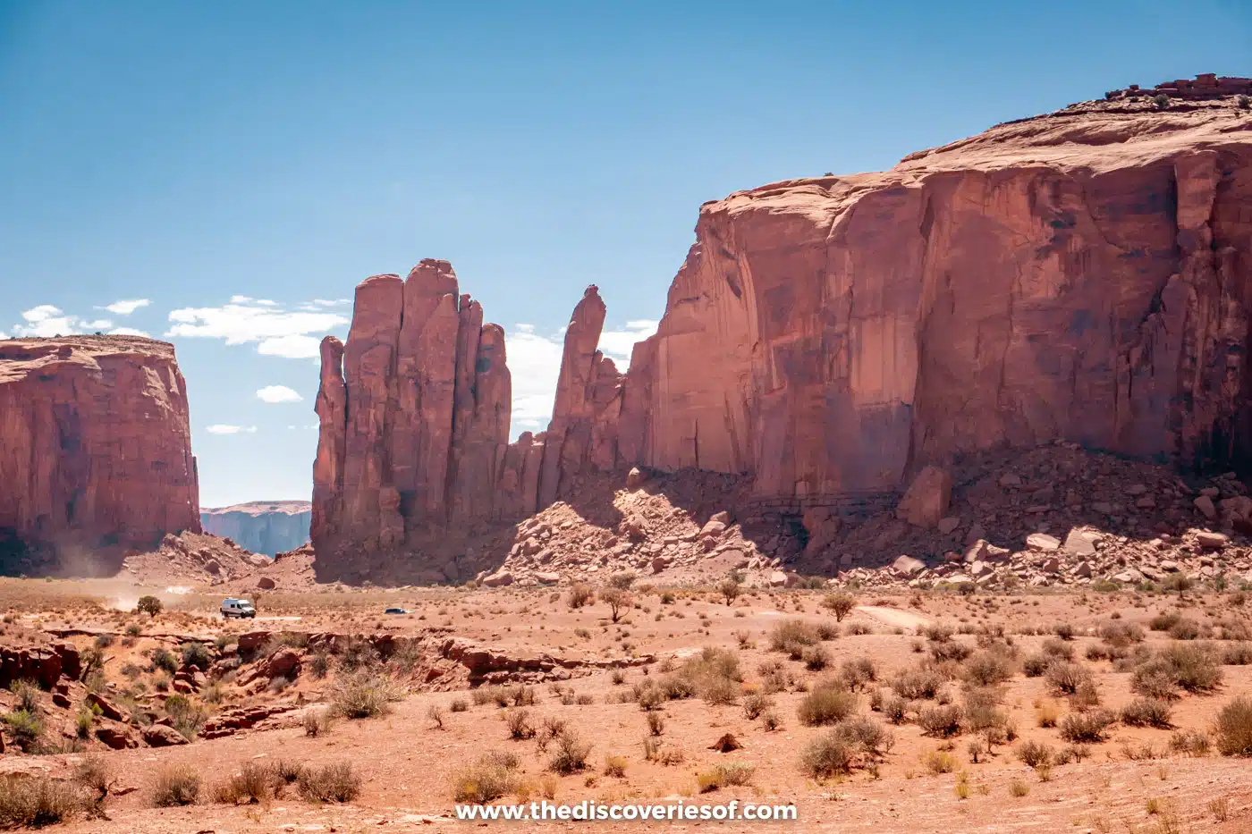 A car dwarfed by towering red buttes on the dirt Valley Drive in Monument Valley