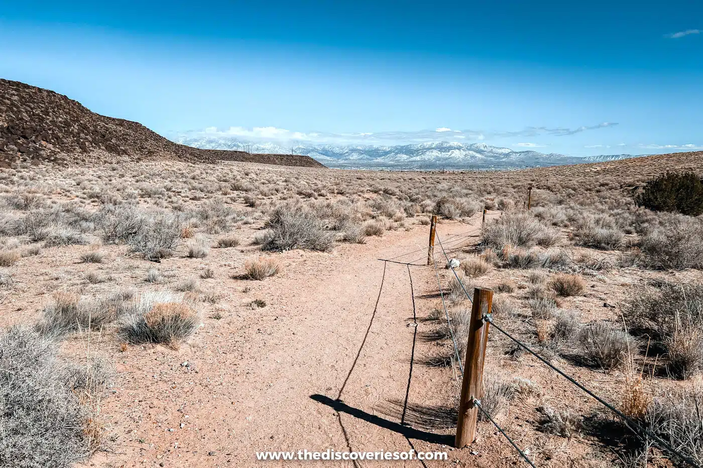 Sandy Rinconada Canyon Trail edged by a rope fence, running through desert scrub towards snowy mountains, Albuquerque