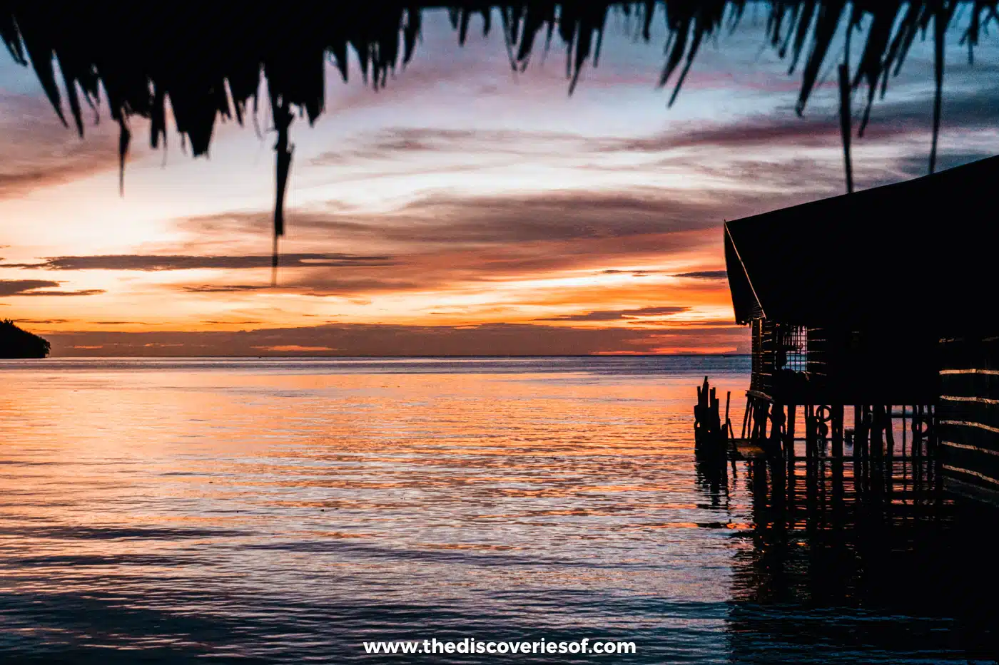 Vivid orange and pink sunset over the sea, framed by thatched eaves and a silhouetted overwater bungalow on stilts