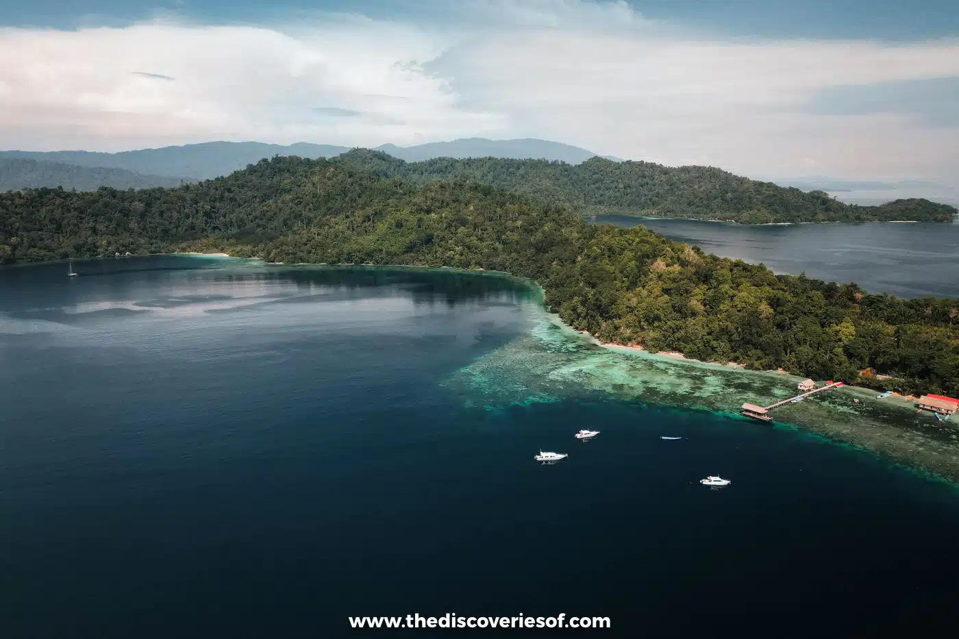Aerial view of Birie Island in Raja Ampat, with boats moored off its reef edge near a wooden jetty and red-roofed buildings