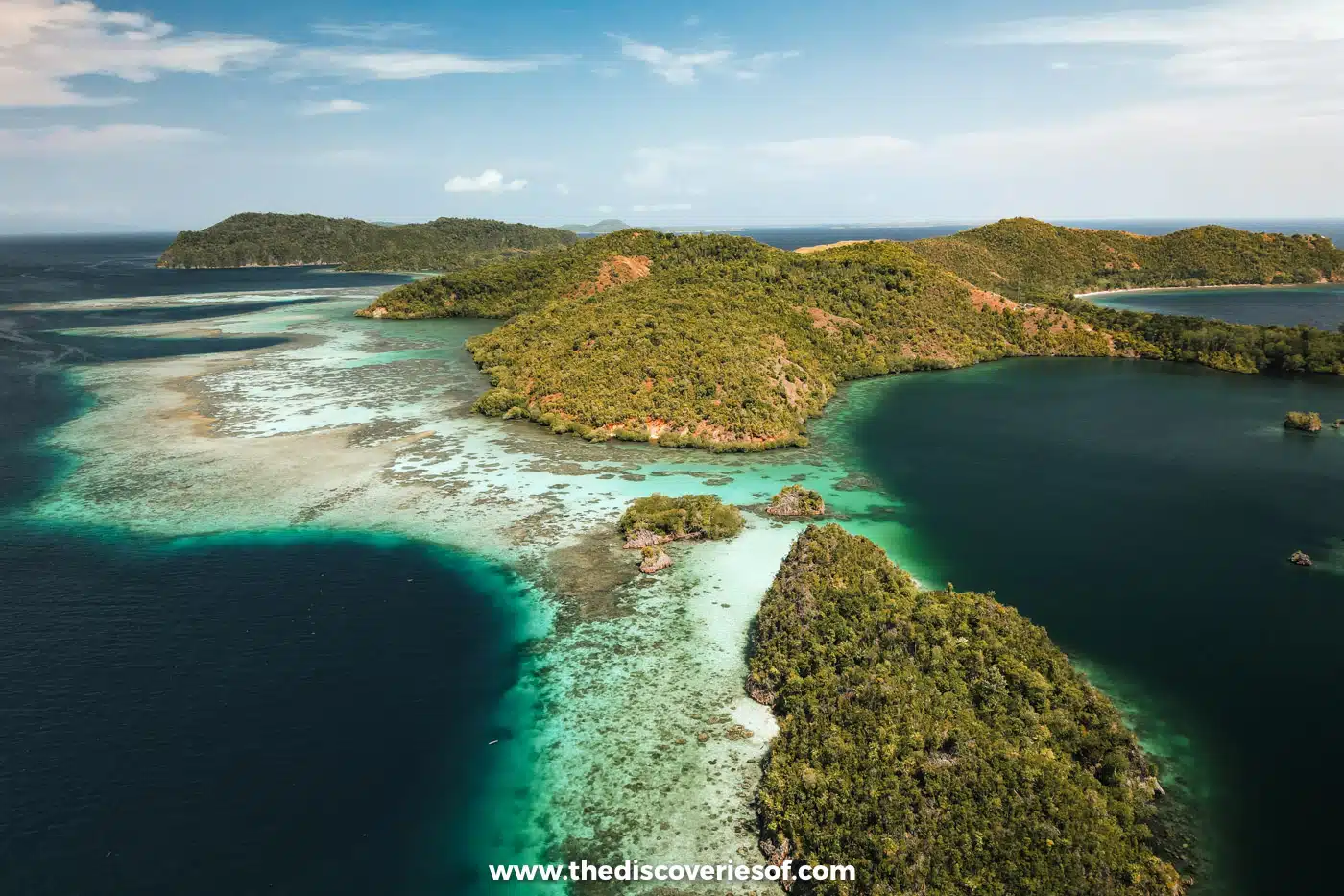 Aerial view of green forested islands ringed by pale reef flats and turquoise shallows dropping into deep blue water