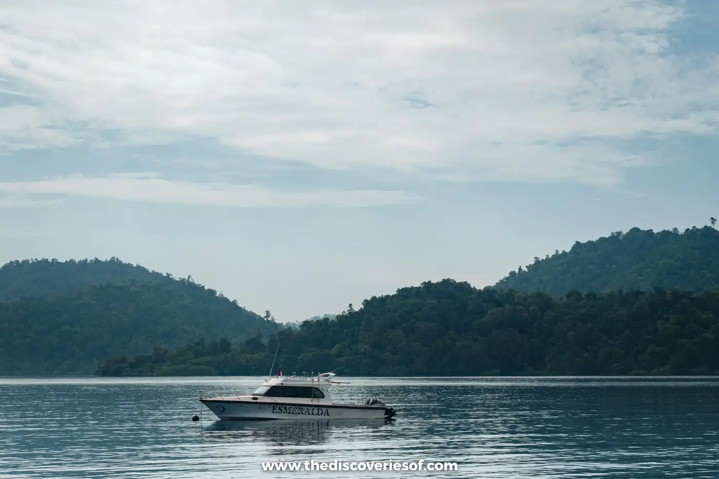 White speedboat moored in a calm bay beneath rolling, forest-covered hills and a soft cloudy sky in Raja Ampat