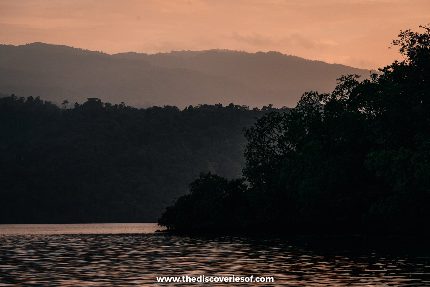 Layered hills and a silhouetted mangrove headland under a peach-coloured sunset sky, reflected in rippled water