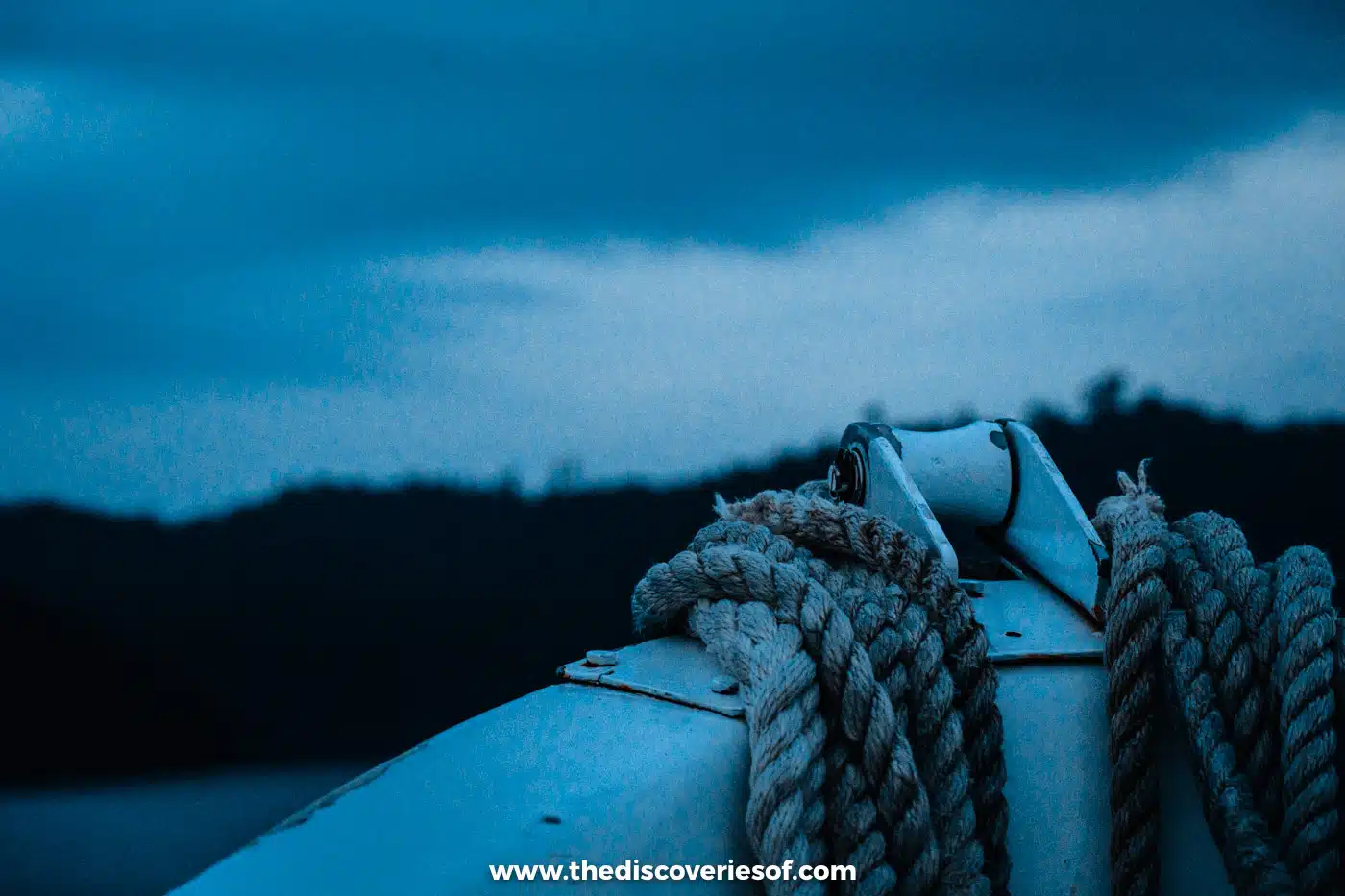 Coiled mooring ropes on the bow of a small boat at dusk, with a dark forested shoreline and cloudy sky behind