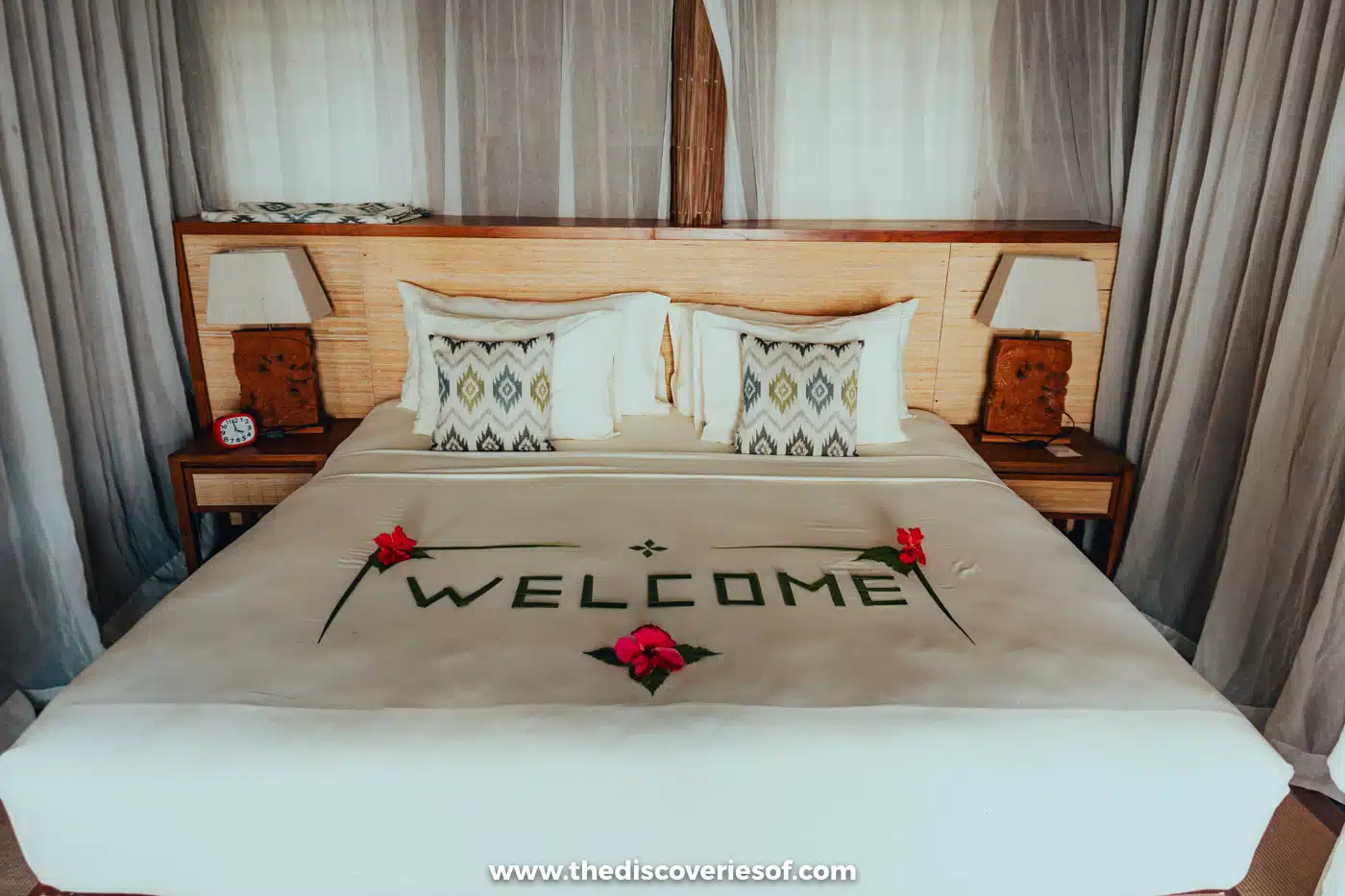 Double bed with patterned cushions, wooden bedside lamps and a welcome in leaves and hibiscus flowers at Papua Paradise