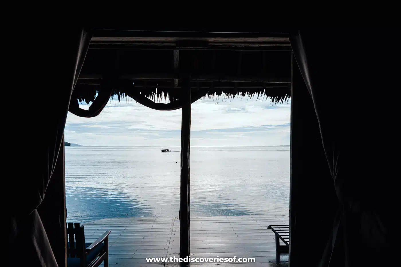 View from inside a darkened bungalow at Papua Paradise through open curtains to a wooden deck, calm sea and a distant boat