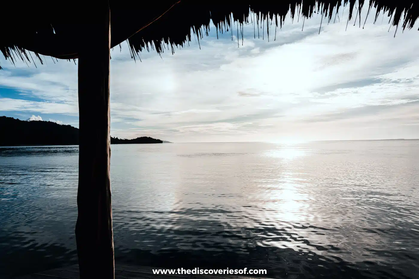 Thatched eave and wooden post framing a calm, silvery sea with sun glare and a low forested island on the left