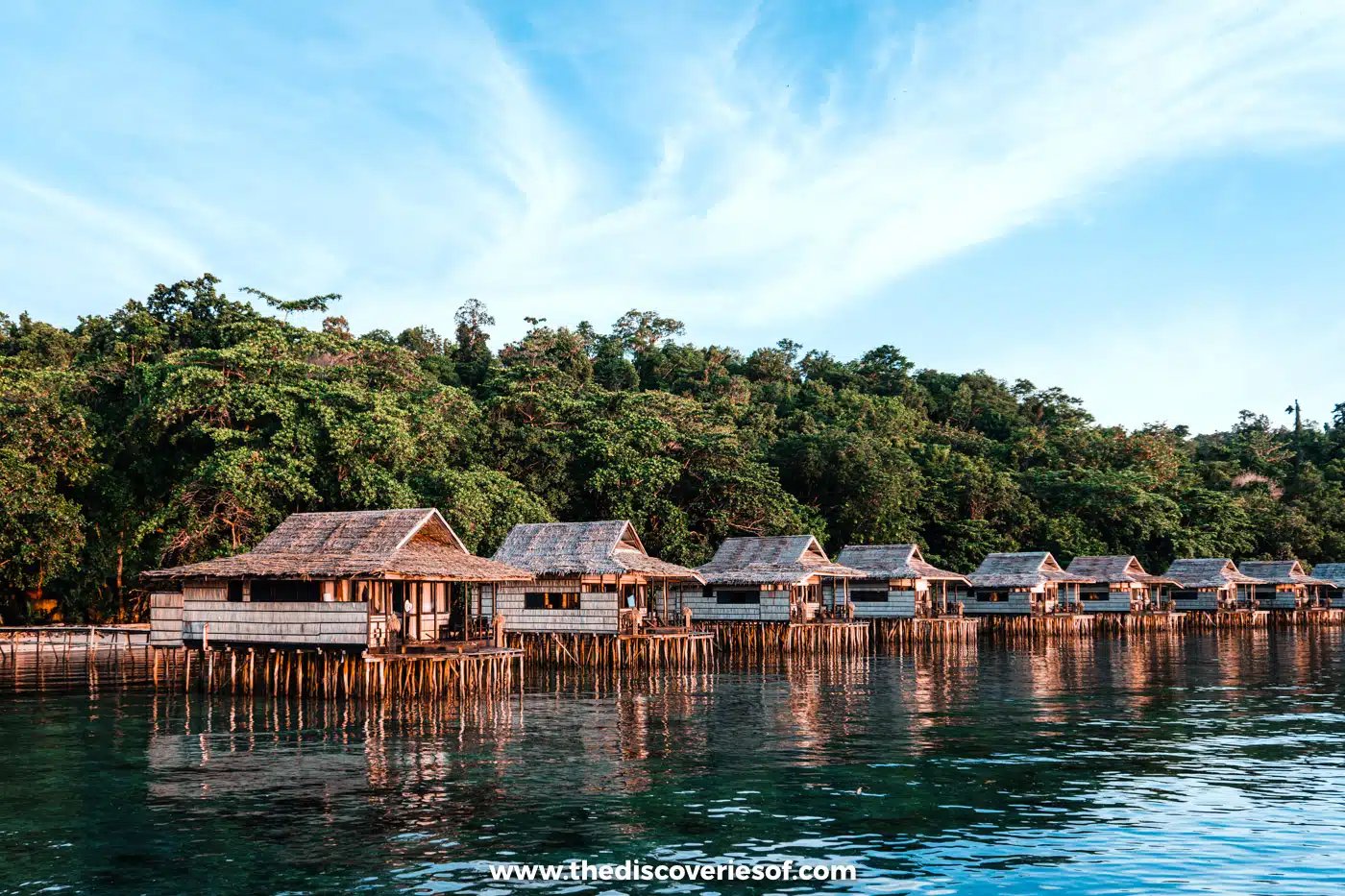 Row of thatched overwater bungalows on wooden stilts above clear green water at Papua Paradise, backed by dense forest