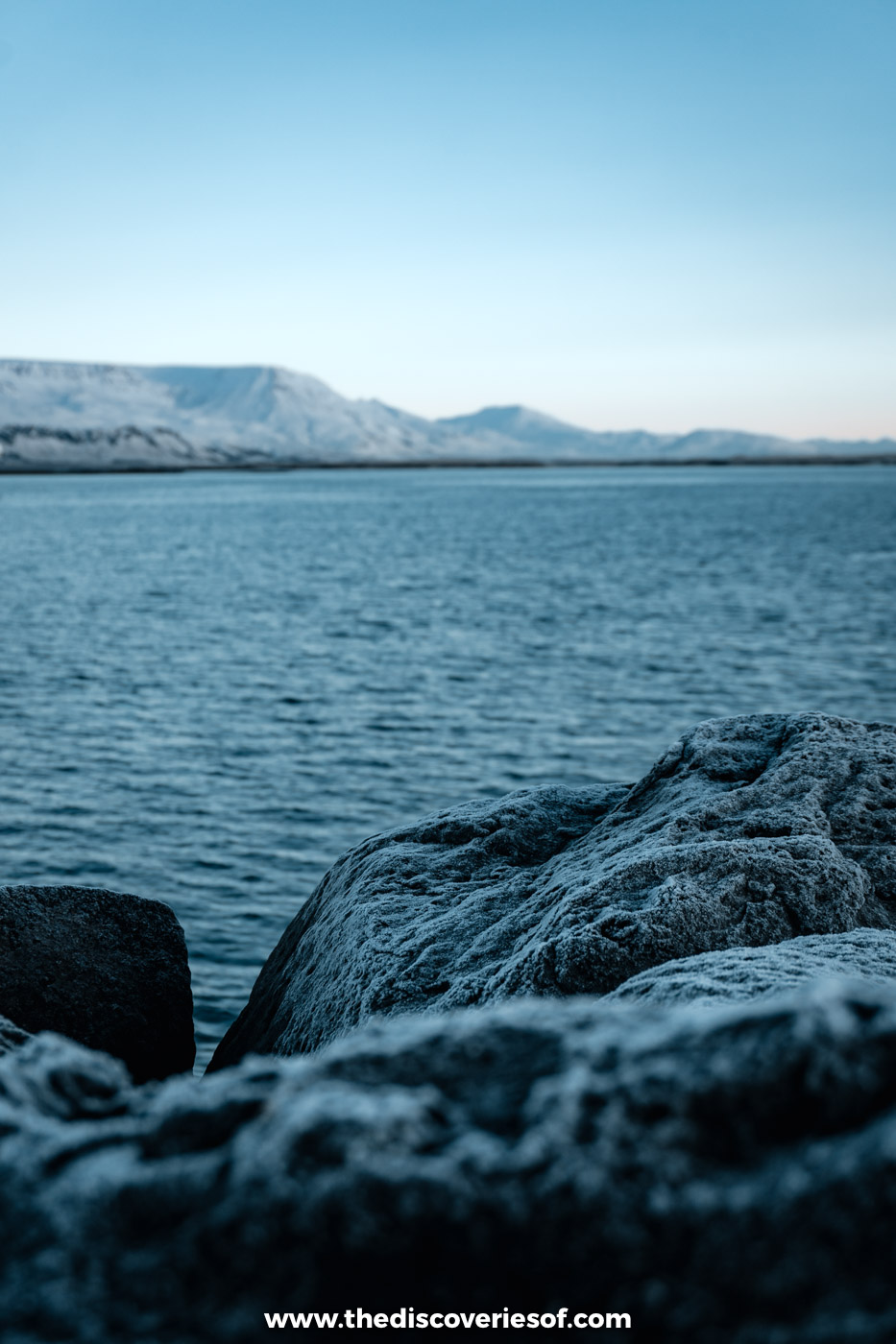 Frosted shoreline rocks and dark water with snow-covered Esja across the bay from Reykjavik in December