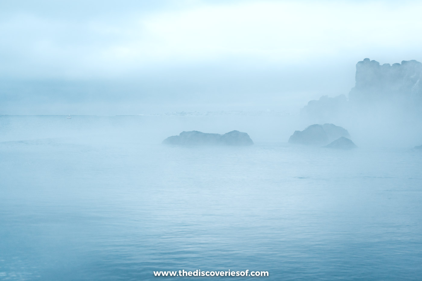 Thick mist drifting over the warm water of Sky Lagoon and the dark sea rocks beyond on a December afternoon