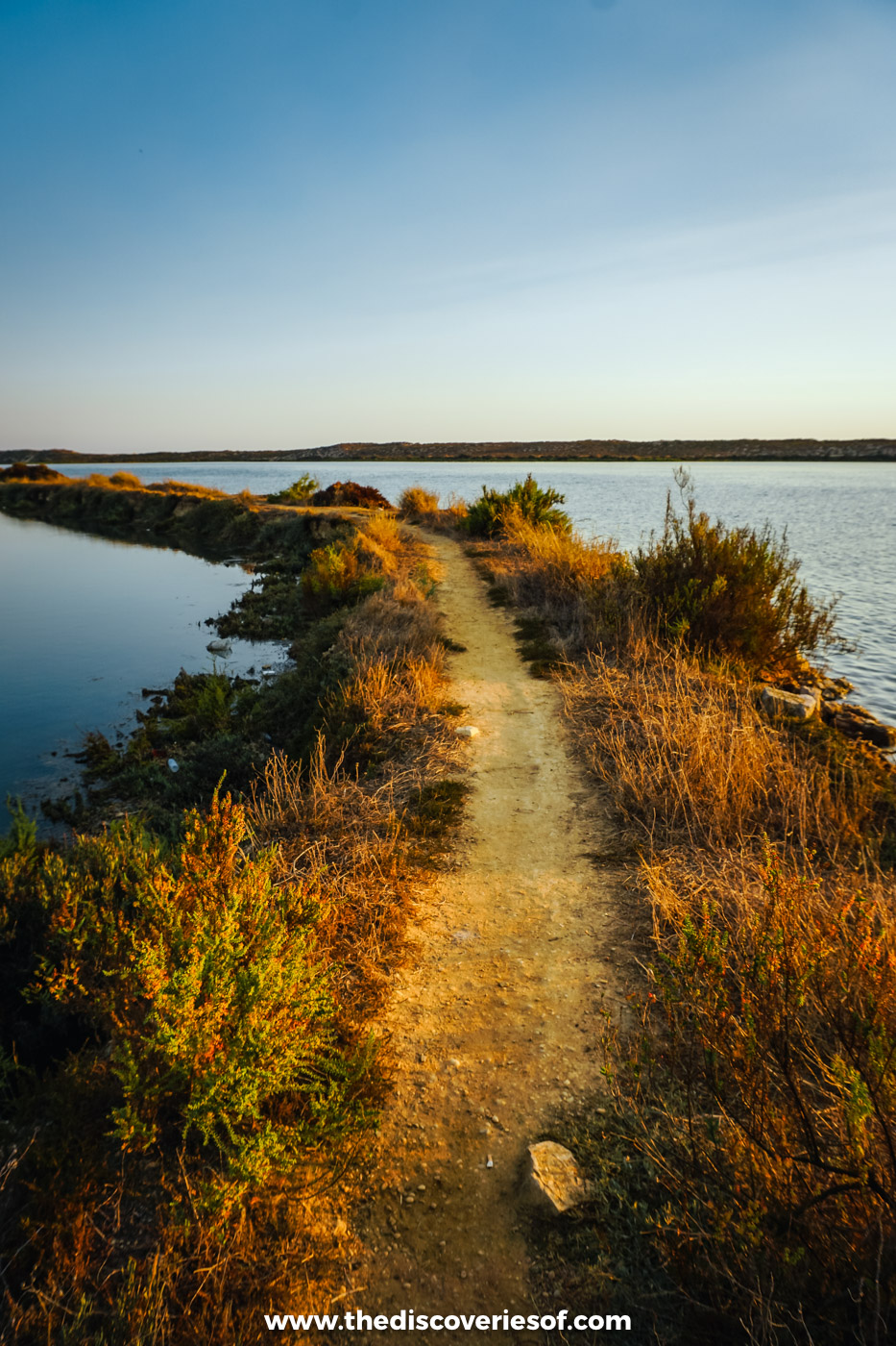 A narrow sandy path running along a strip of golden scrub between two stretches of still lagoon water near Quinta do Lago