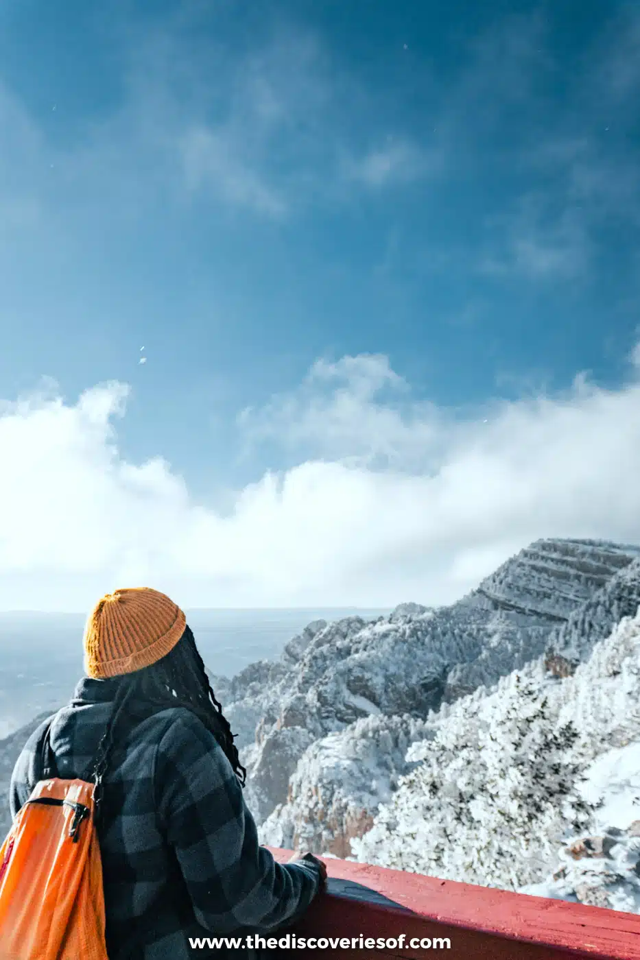 Julianna in an orange beanie and backpack looking out over the snowy Sandia Mountains from the top of the tram
